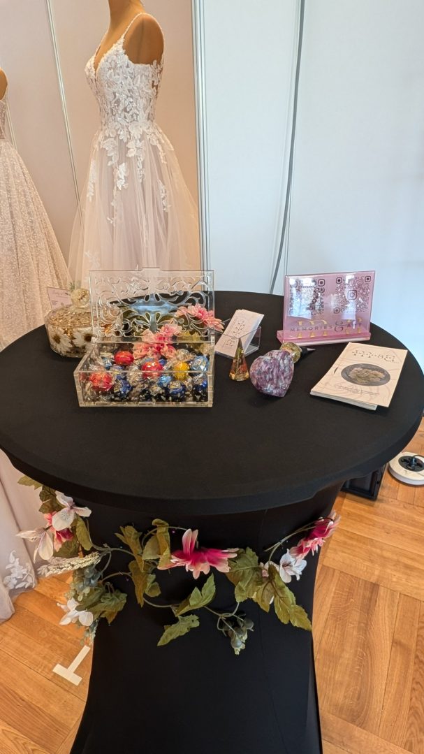 Runder Tisch mit Dekoration, Blumen und Brautkleidern im Hintergrund.